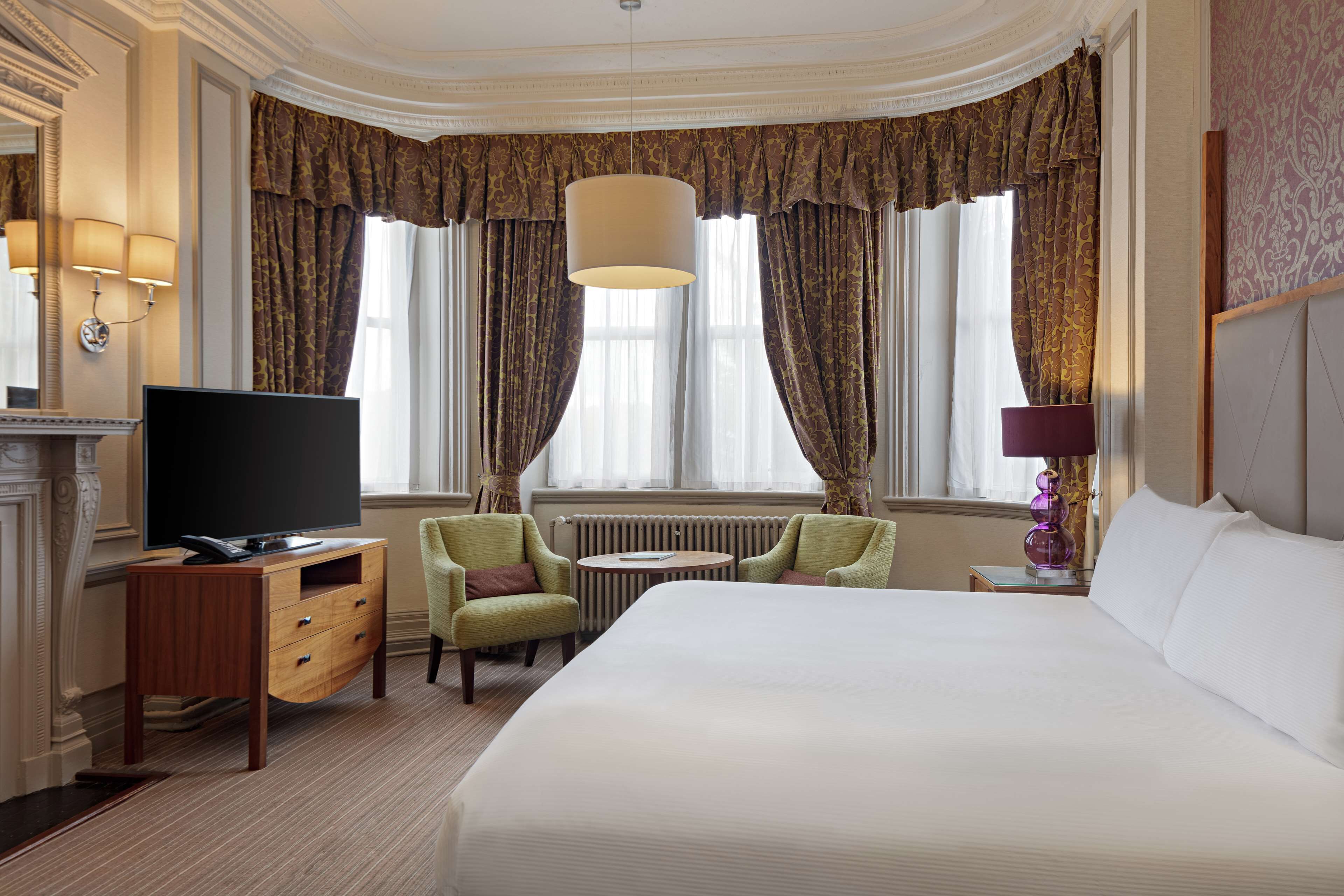 A hotel room with a large bed, a television on a wooden dresser, two armchairs, a small round table, and a fireplace.
