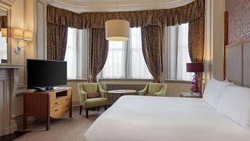 A hotel room with a large bed, a television on a wooden dresser, two armchairs, a small round table, and a fireplace.