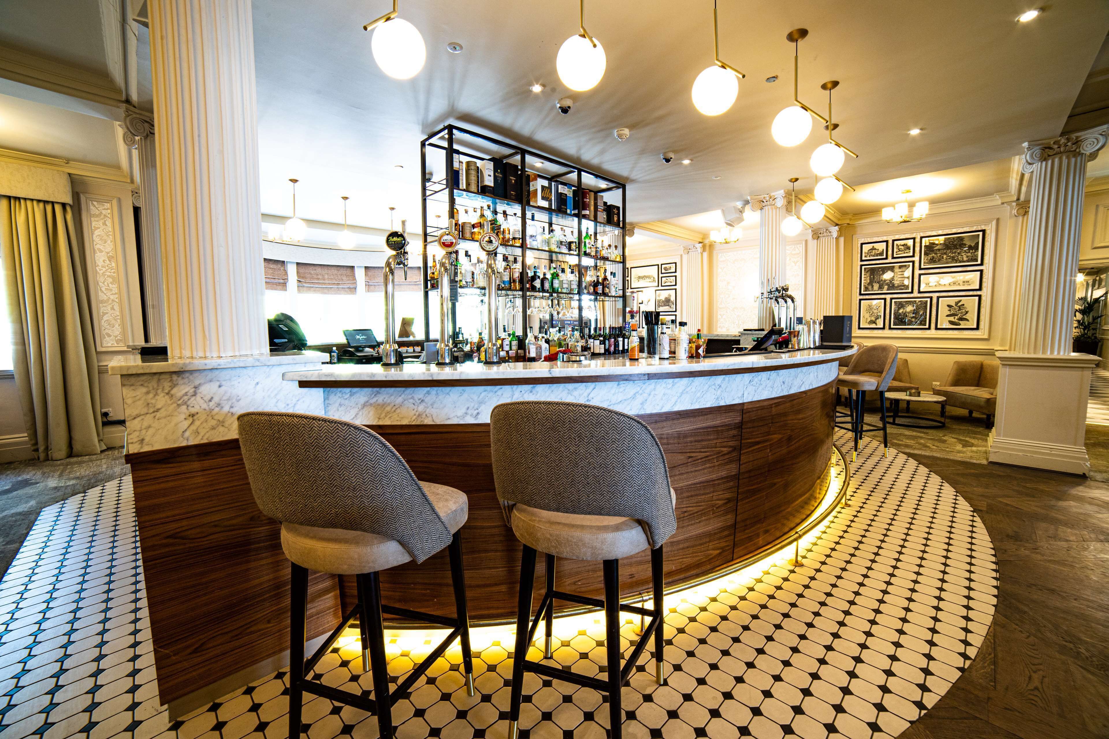 A bar area with a marble countertop, two bar stools, a shelf with bottles, and a patterned floor.