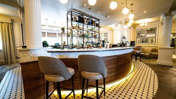 A bar area with a marble countertop, two bar stools, a shelf with bottles, and a patterned floor.