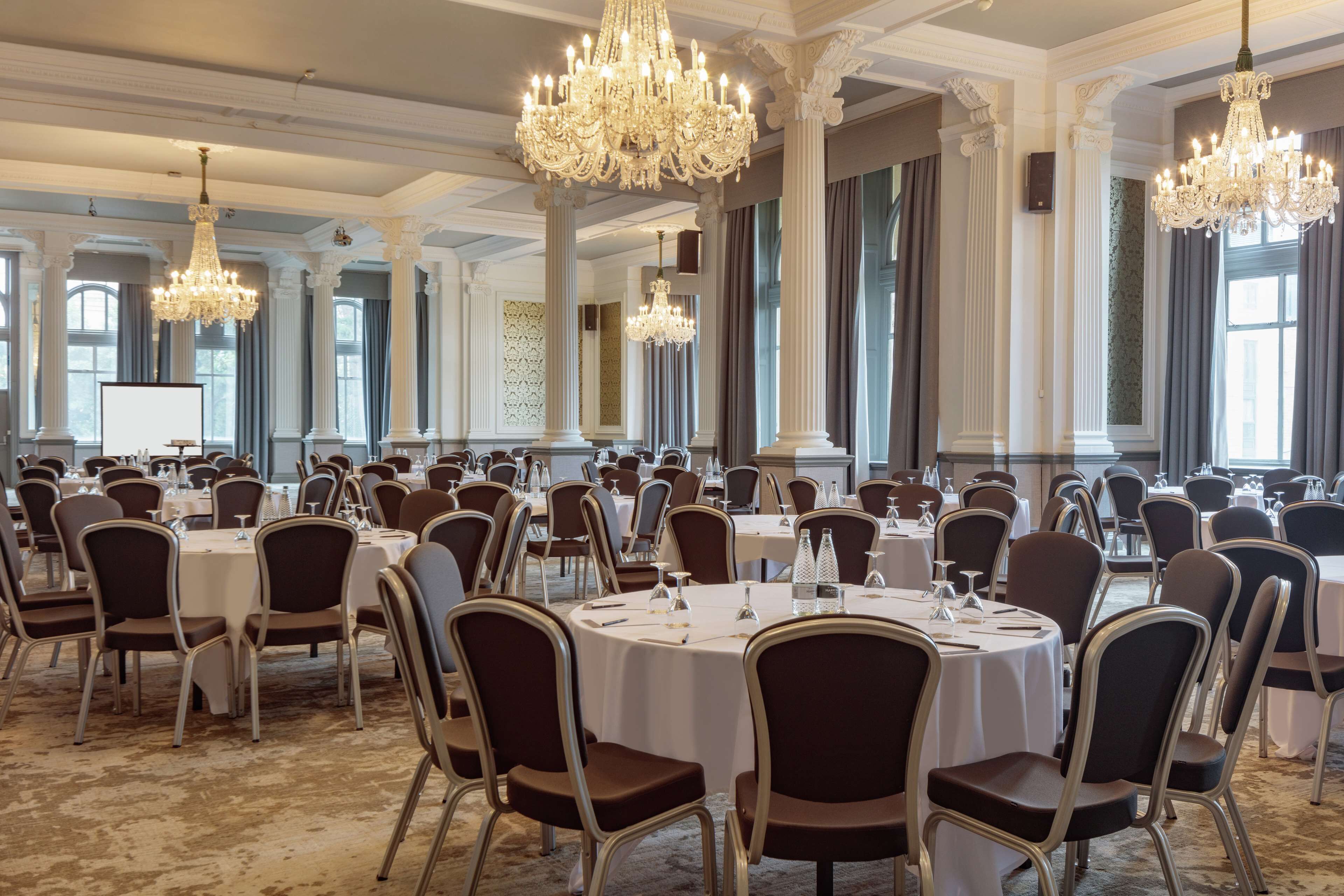 Een feestzaal met ronde tafels, stoelen, kroonluchters en zuilen.
