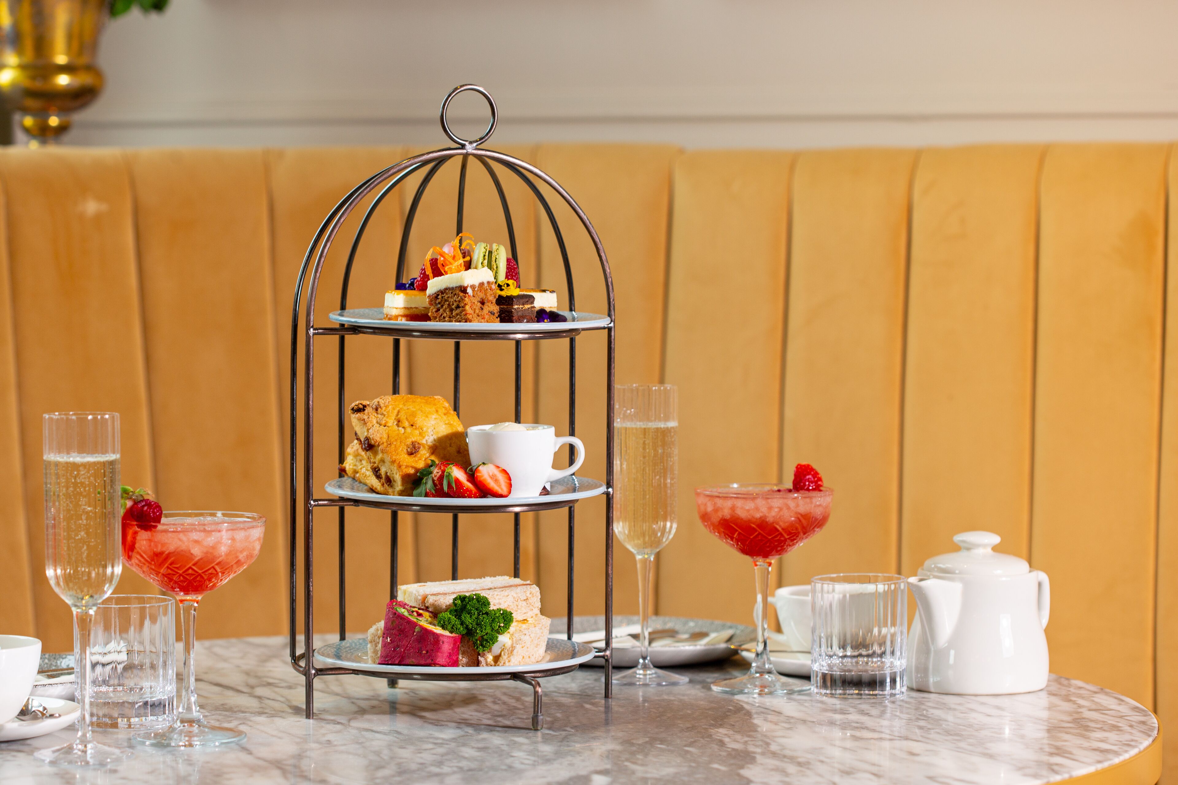 A three-tiered dessert stand with various pastries and sandwiches, accompanied by drinks and a teapot on a marble table.