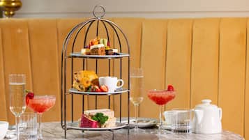 Un soporte de tres niveles con diversos pasteles y sándwiches, acompañado de bebidas y una tetera sobre una mesa de mármol.