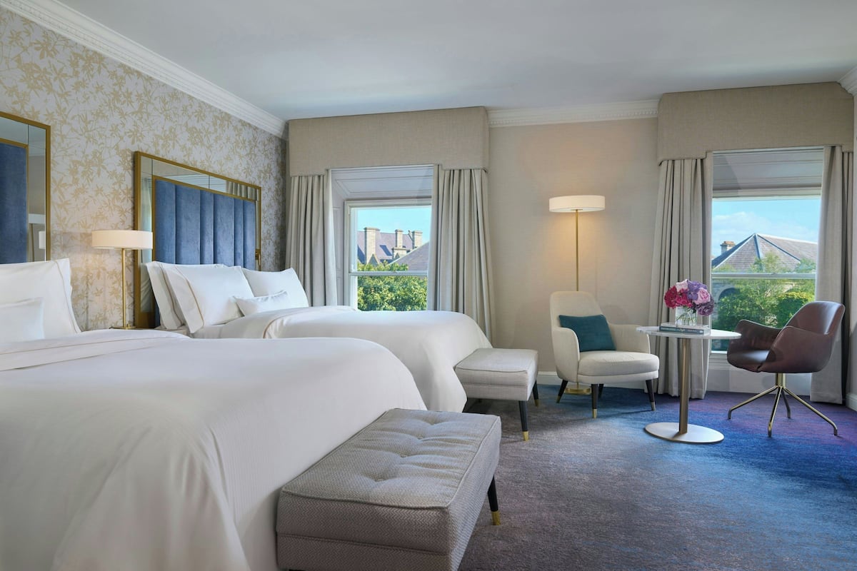 Habitación de hotel con dos camas, una zona de estar con una silla y una mesa, y vistas al exterior.