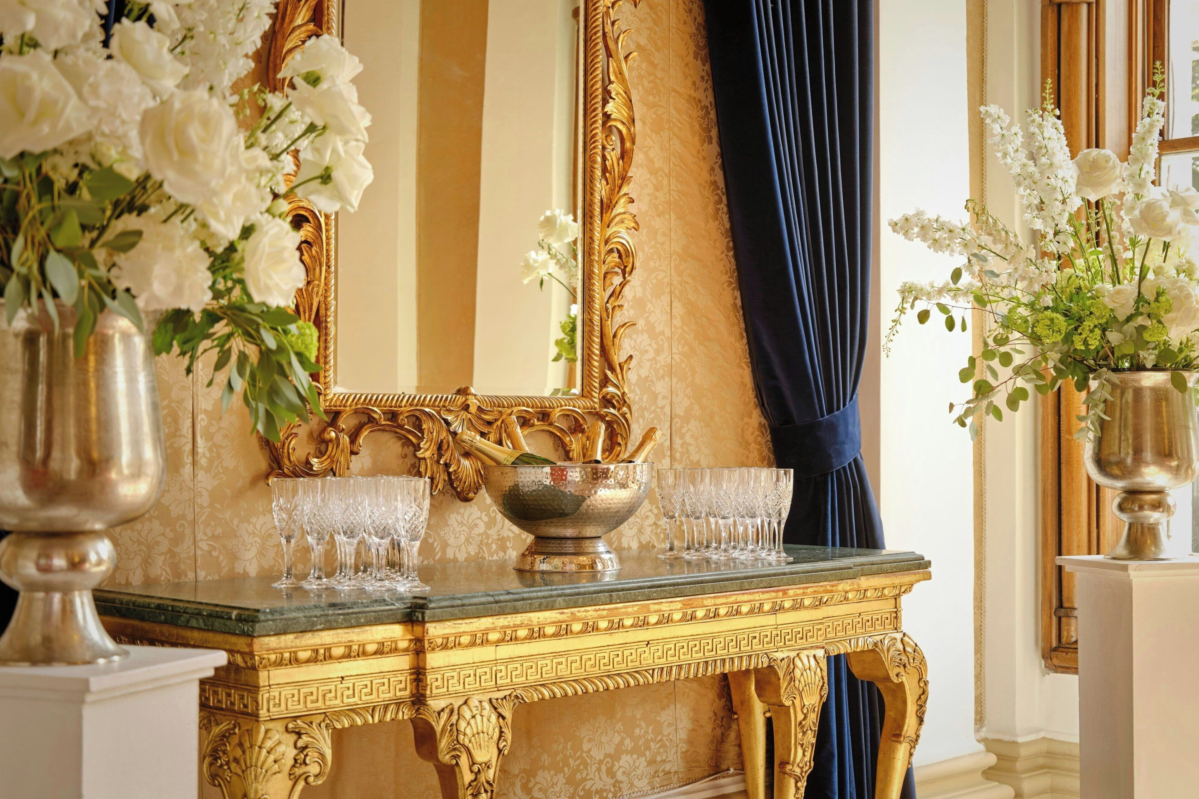 A luxurious room with a gold-framed mirror, a marble table with a gold decorative border, and a vase of white flowers.