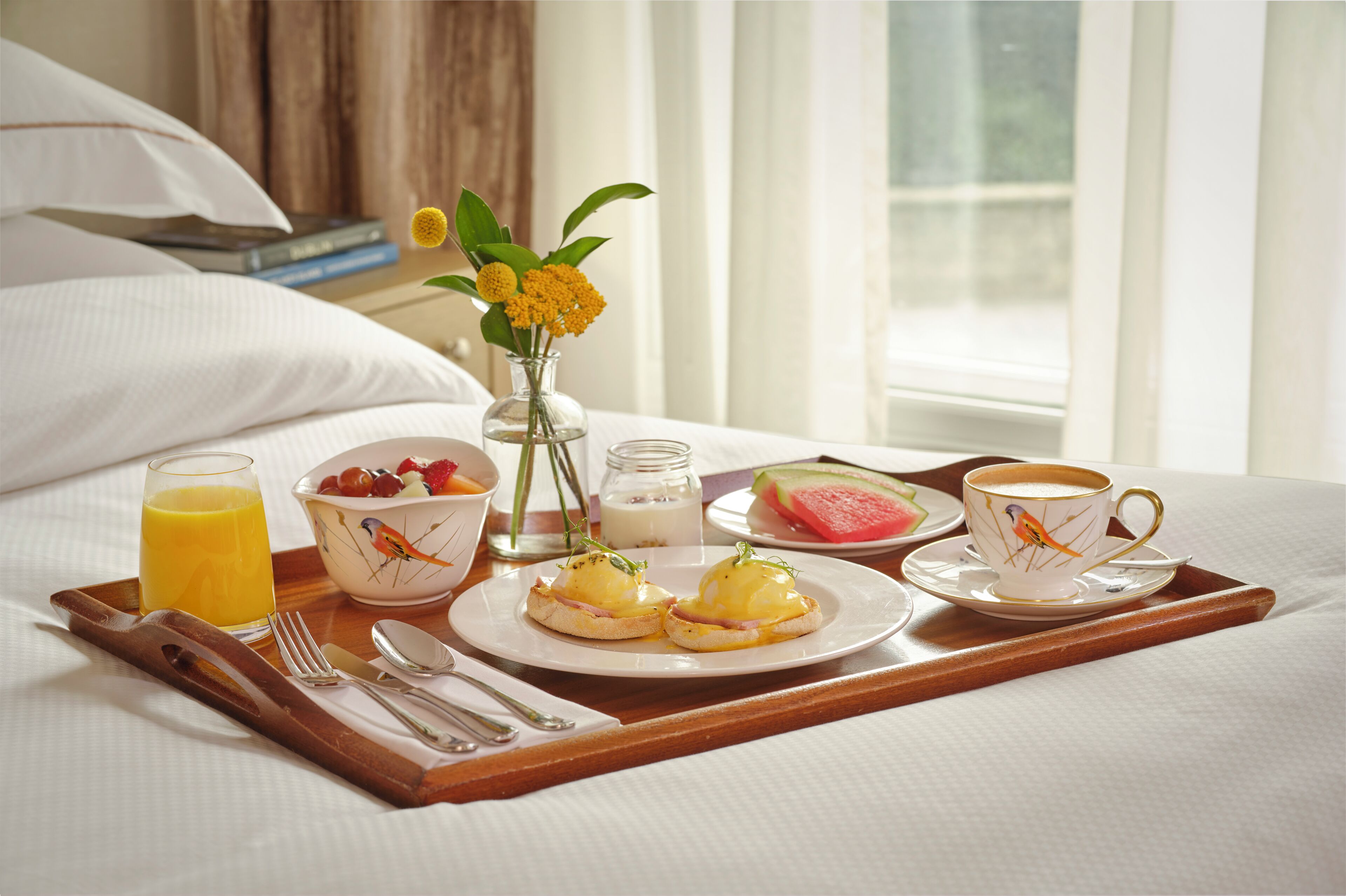 A breakfast tray with eggs benedict, a glass of orange juice, a bowl of fruit, a cup of coffee, and a vase with flowers on a bed.