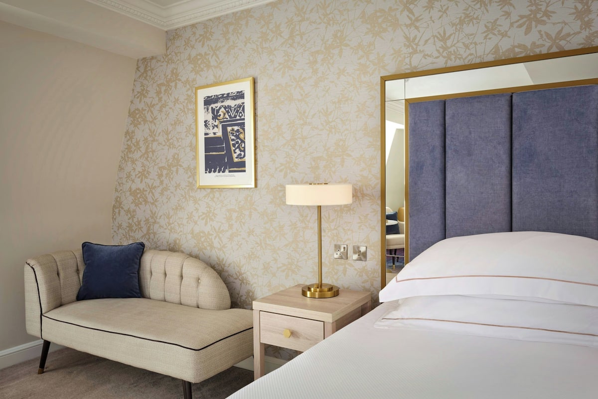 Habitación de hotel con cama, mesita de noche con lámpara, sofá e imagen enmarcada en la pared.