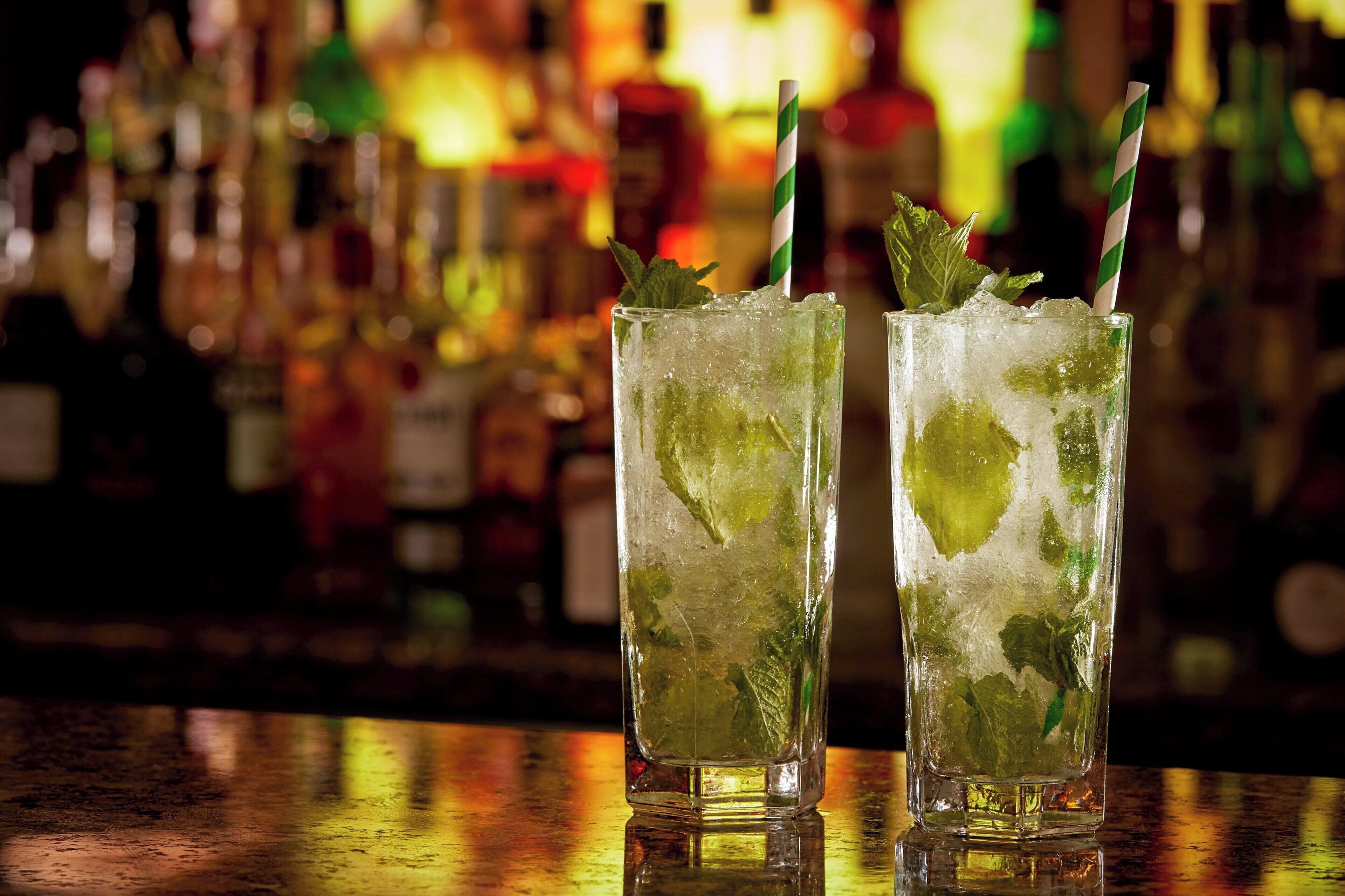 Deux grands verres contenant un cocktail vert, avec des glaçons et des feuilles de menthe, et des pailles rayées, posés sur le comptoir d’un bar.