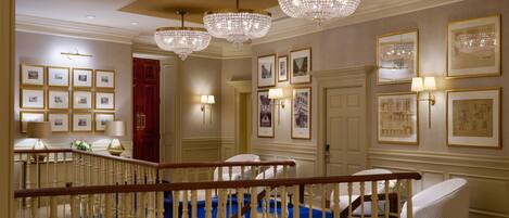 Un amplio pasillo con candelabros, cuadros enmarcados y una escalera tapizada en azul.