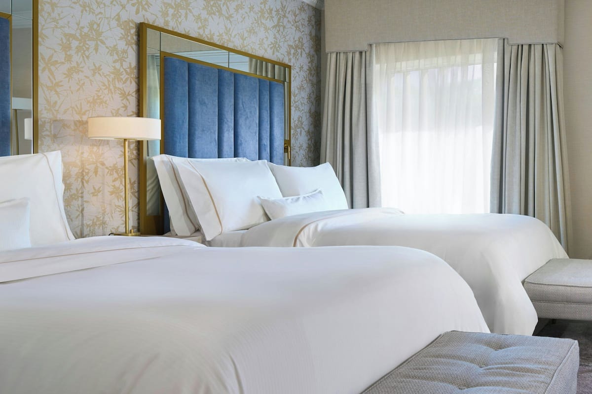 Habitación de hotel con dos camas, un espejo grande, una lámpara y vistas al exterior a través de cortinas translúcidas.