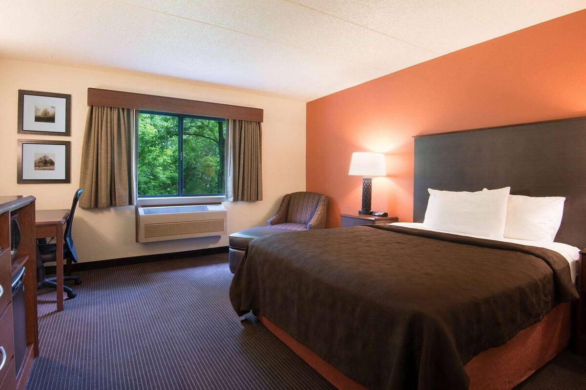 Ein Hotelzimmer mit Bett, Schreibtisch, Stuhl, Fernseher, einem Fenster mit Vorhängen und einer Lampe.