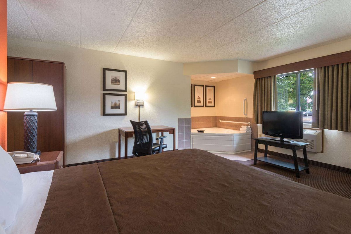 Ein Hotelzimmer mit einem großen Bett, einem Schreibtisch mit Stuhl, einem Fernseher, einer Badewanne und einem Fenster mit Vorhängen.