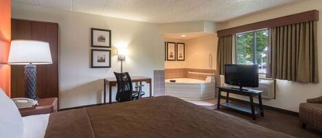 A hotel room with a large bed, a desk with a chair, a television, a bathtub, and a window with curtains.