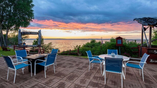 An outdoor patio with a table and chairs, overlooking a body of water at sunset. - AmericInn by Wyndham Ashland (Ashland)