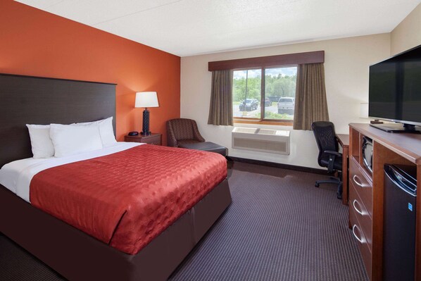 A hotel room with a large bed, a desk with a television, a chair, a window with curtains, and a heater. - AmericInn by Wyndham Ashland (Ashland)