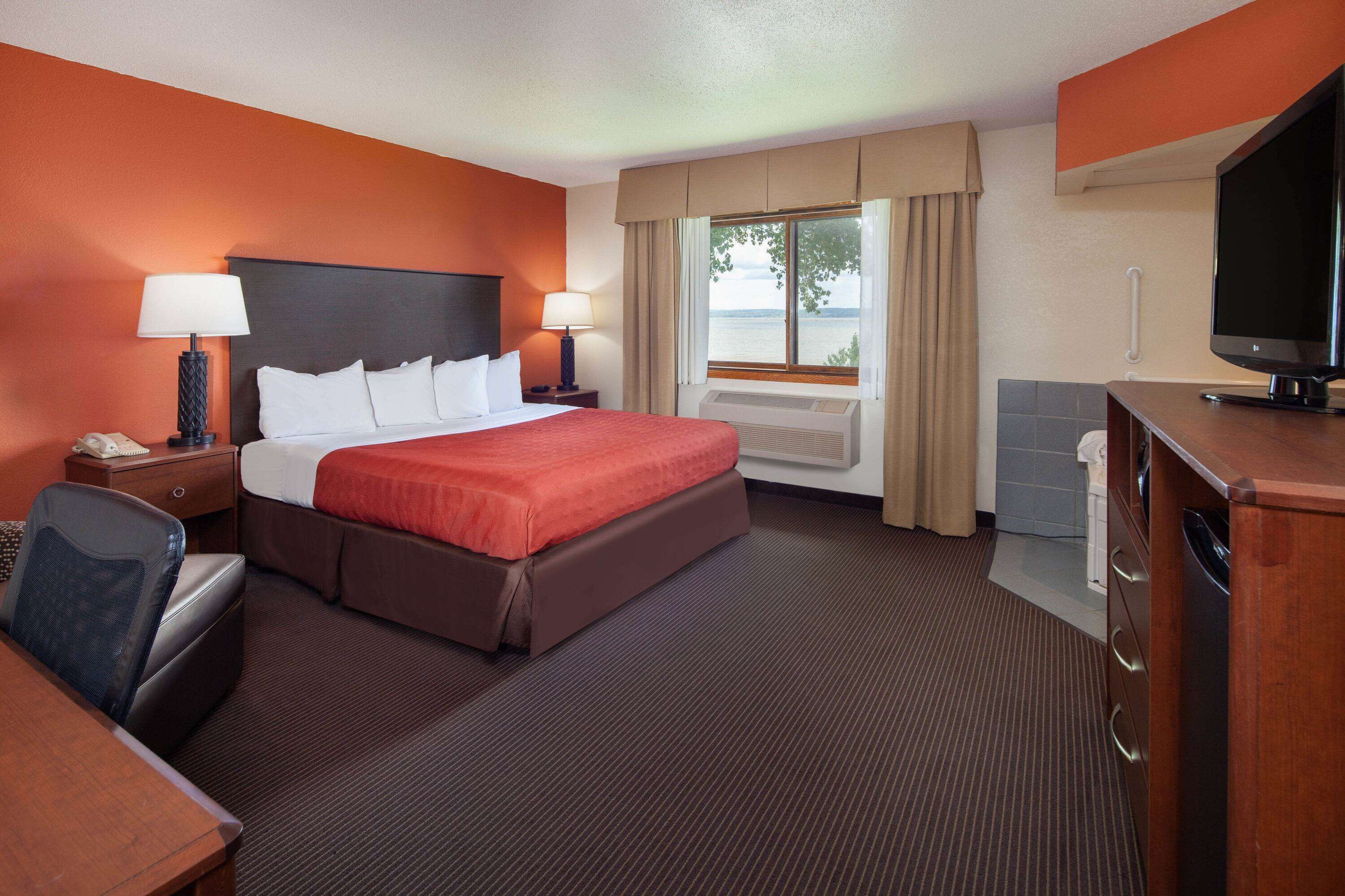 A hotel room with a large bed, two bedside tables with lamps, a desk with a chair, a television, and a view of the outdoors through a window.