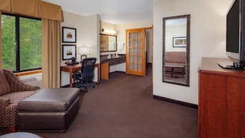 A hotel room with a large window, a sofa, a desk with a computer, a television, and a bathroom with a mirror and sink.