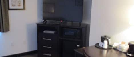 A small, dimly lit room with a dark wooden desk, a chair, a television mounted on the wall, and a framed picture on the wall.