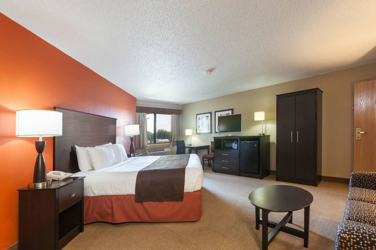 A hotel room with a bed, bedside table, lamp, desk, chair, TV, wardrobe, and a small round table.