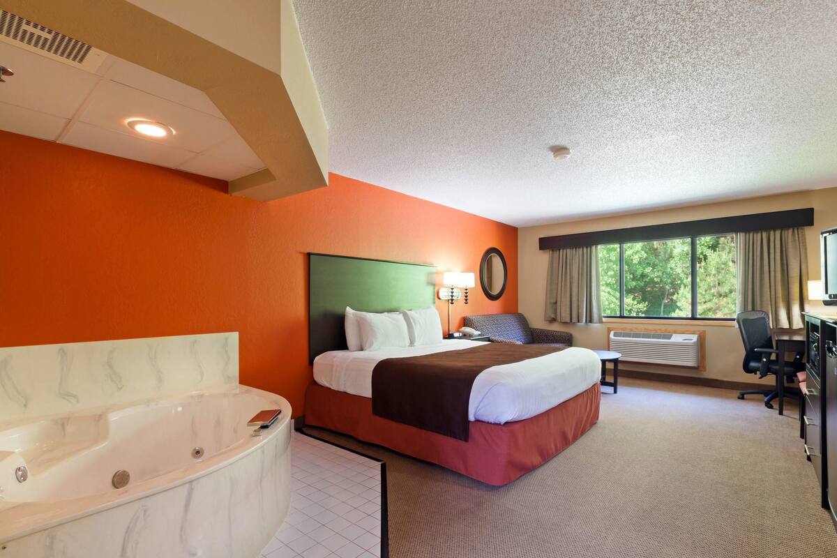 A hotel room with a large bed, a separate bathroom with a jacuzzi, a desk, and a window with curtains.