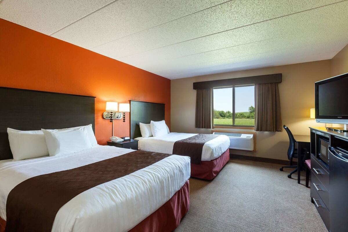 A hotel room with two beds, a desk with a television, a window with a view, and an air conditioning unit.