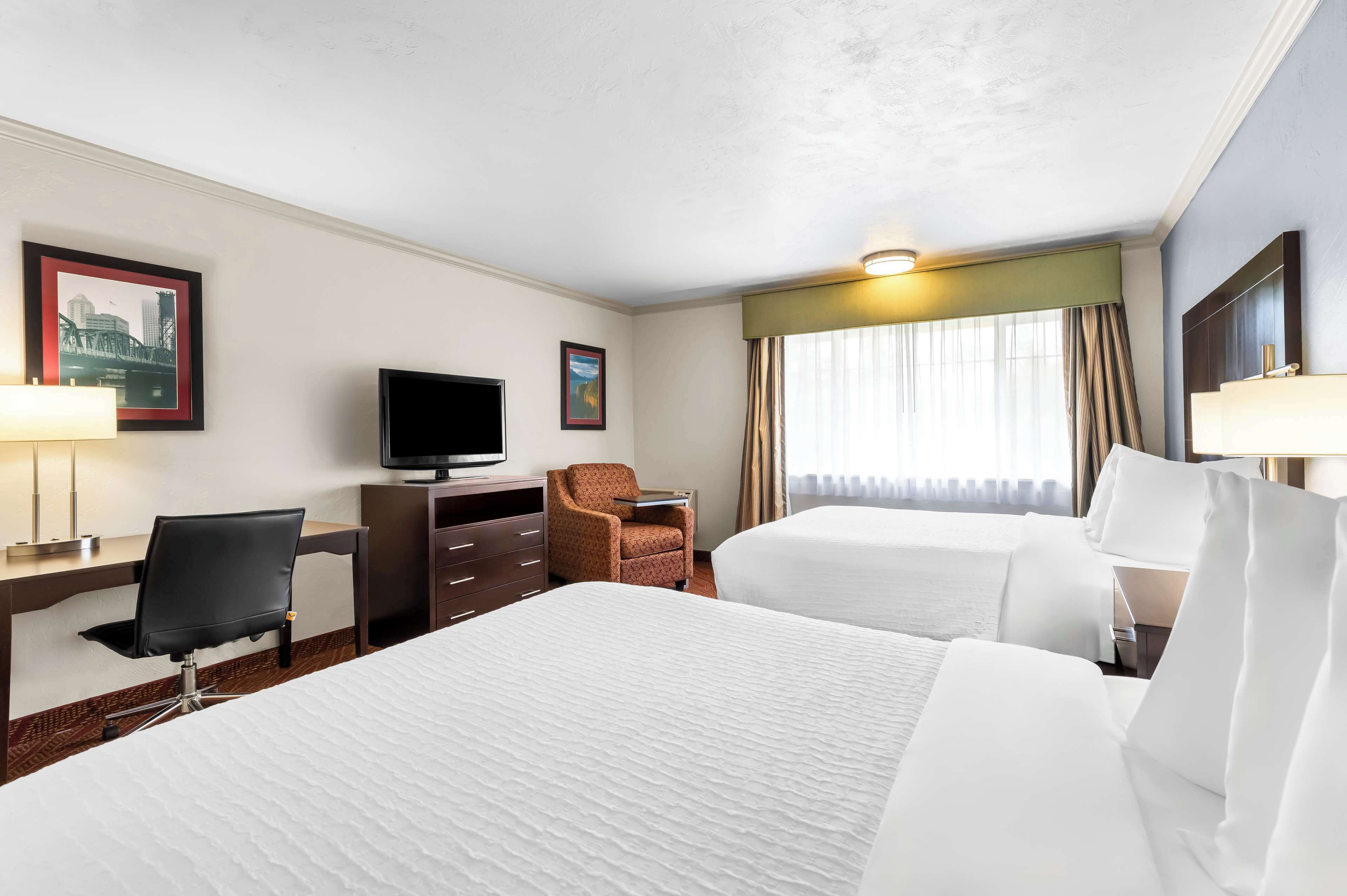 Ein Hotelzimmer mit zwei Betten, einem Schreibtisch mit Stuhl, einem Fernseher und einem Fenster mit Vorhängen.