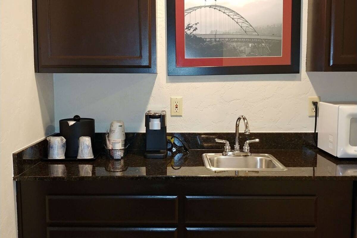 A kitchen with dark wooden cabinets, a stainless steel sink, a coffee maker, and a framed picture of a bridge on the wall.