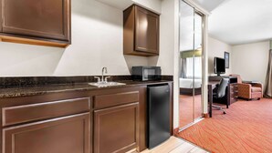 A compact kitchen with dark brown cabinets, a black refrigerator, and a microwave. There is a small sink and a glass door leading to a living area with a sofa and a television. - Best Western Plus Parkway Inn (Wilsonville)