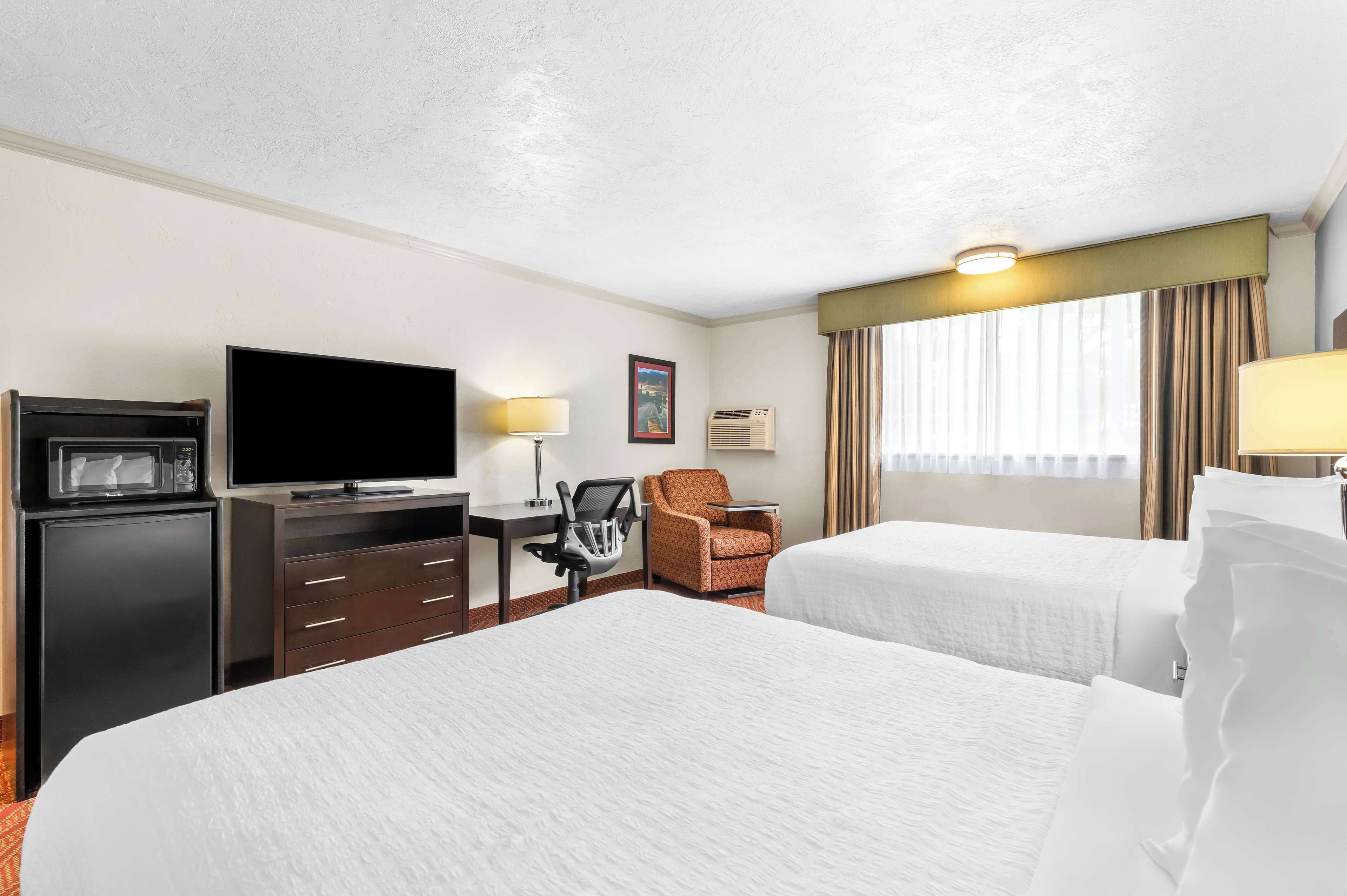 A hotel room with two beds, a television, a desk with a chair, and a window with curtains.