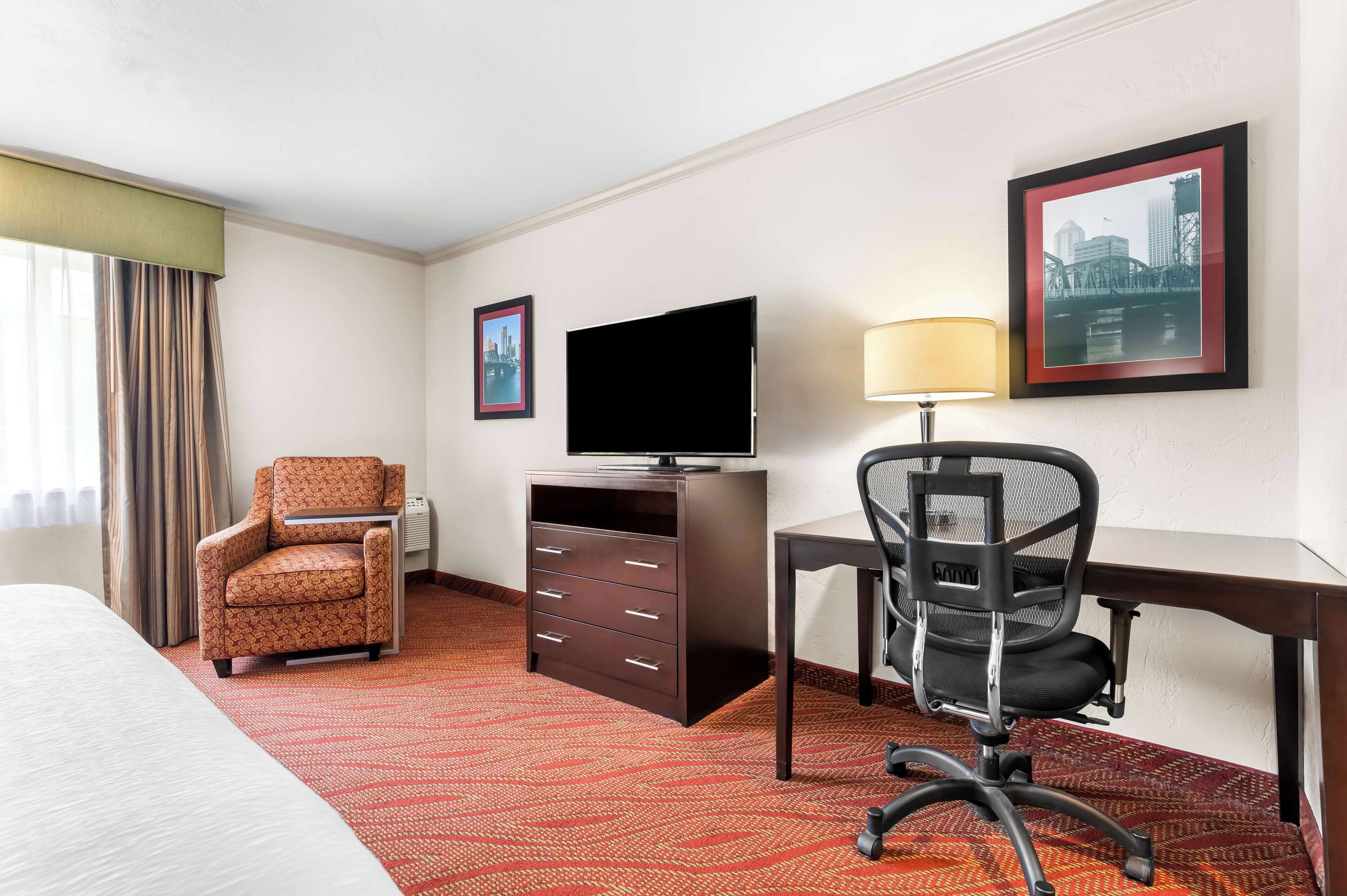 Une chambre d’hôtel avec un lit, un bureau, une chaise, un téléviseur et un tableau représentant un paysage urbain accroché au mur.