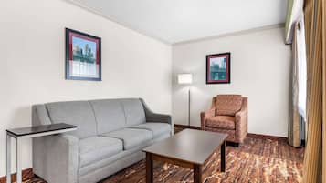 A hotel room with a grey sofa, a patterned armchair, a small table, a floor lamp, and two framed pictures on the walls.
