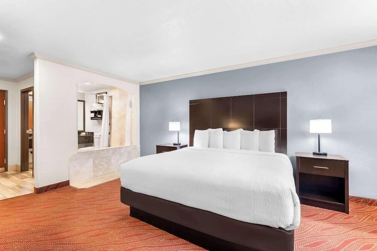 A hotel room with a large bed, two bedside tables with lamps, a mirror, and a bathroom visible through an open door.