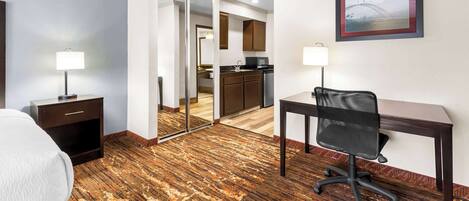 A hotel room with a bed, desk, chair, and a view of the kitchen through a glass door.