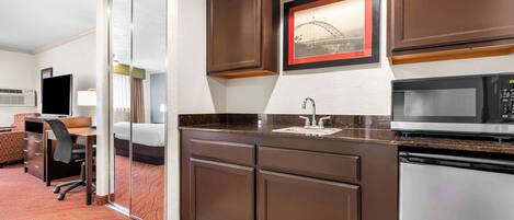 Cocina de hotel compacta con microondas, lavamanos y muebles oscuros.