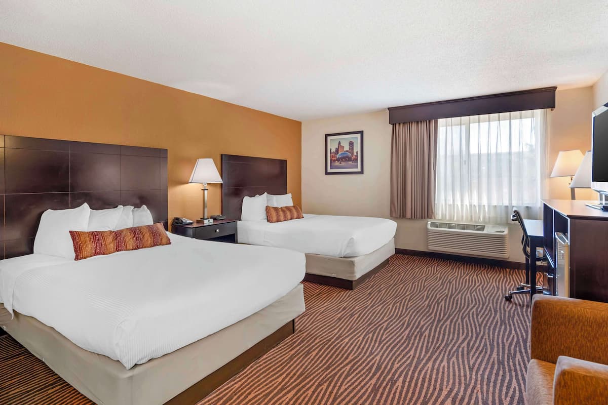 A hotel room with two beds, a desk, a chair, a window with curtains, and a wall-mounted air conditioner.