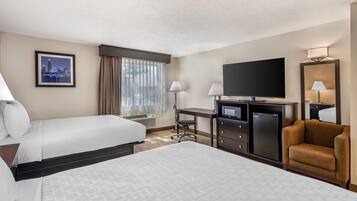 A hotel room with two beds, a desk with a flat-screen TV, a minibar, and a seating area with a chair.