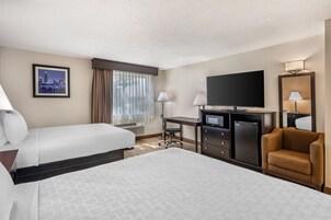 Habitación de hotel con dos camas, un escritorio, un televisor de pantalla plana, un minibar y una zona de estar con una silla.