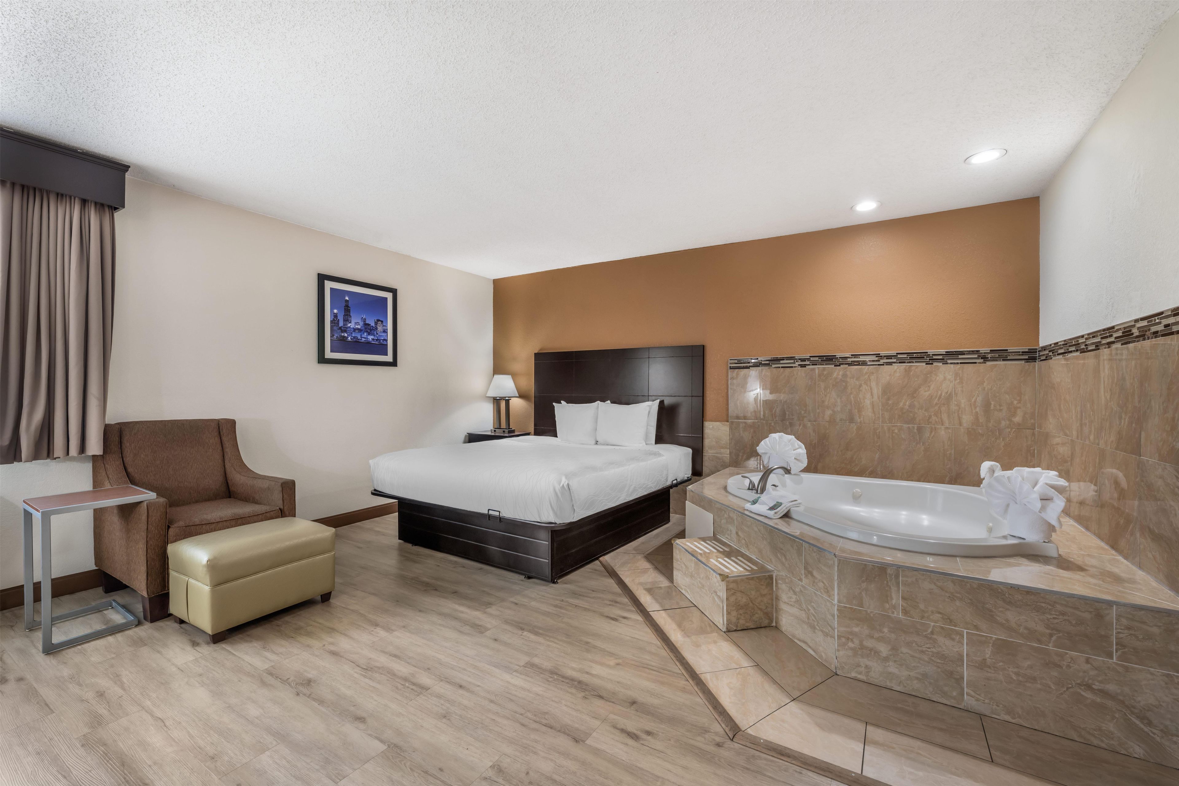 A hotel room with a bed, a chair, a small table, a bathroom with a bathtub, and a framed picture on the wall.