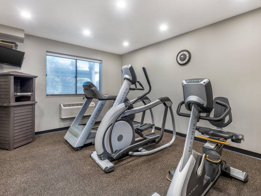 Een sportschool met loopbanden, een elliptische trainer en een hometrainer, een televisie en een klok aan de muur.