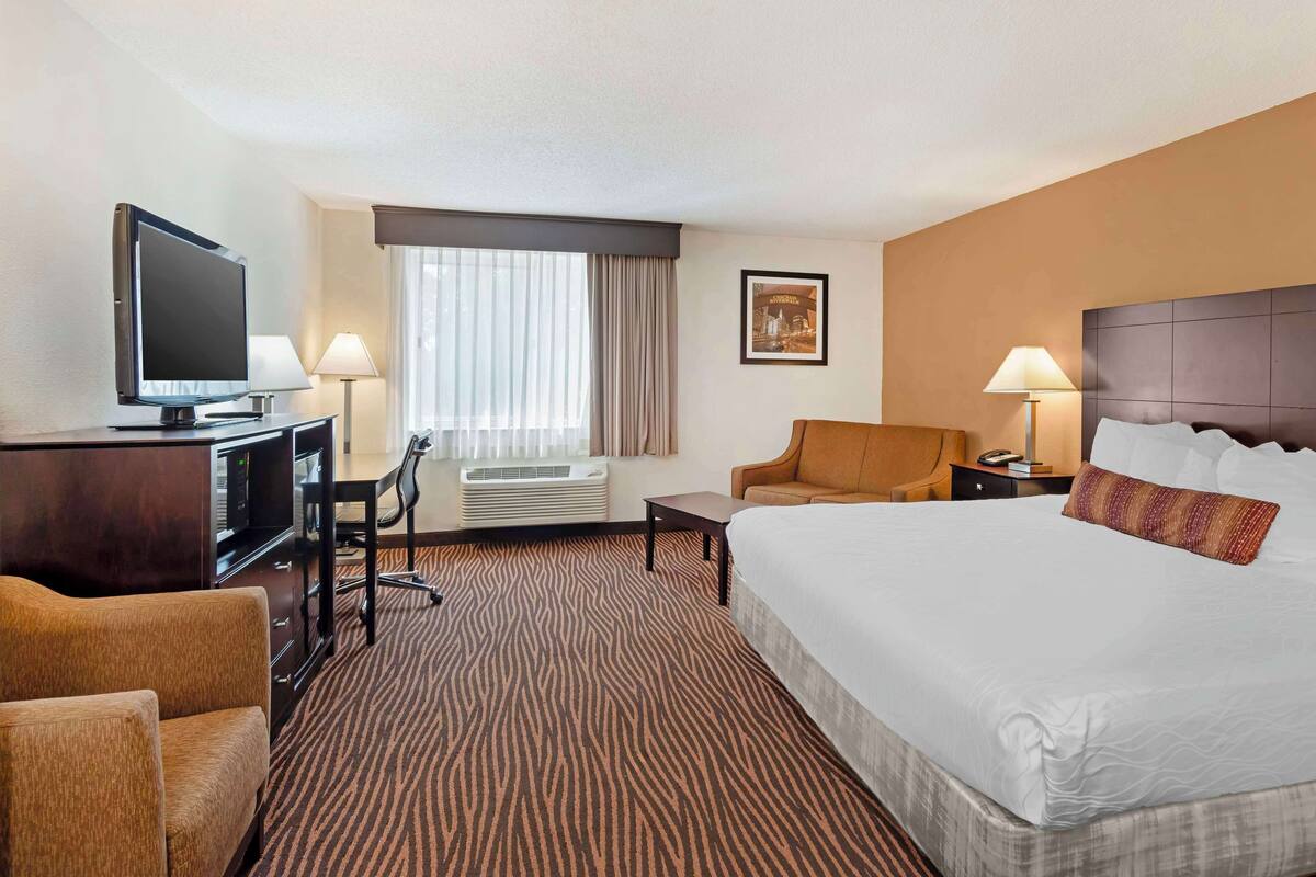 A hotel room with a large bed, a desk with a computer, a television, a chair, a lamp, and a window with curtains.