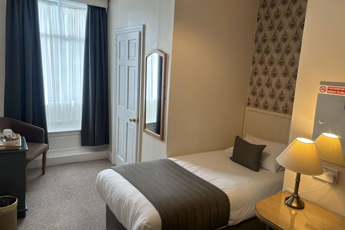 A hotel room with a bed, bedside table with lamps, a desk with a chair, a mirror, and a window with curtains.