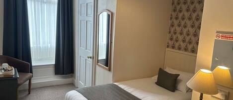 Habitación de hotel con cama, mesita de noche con lámparas, escritorio con silla, espejo y ventana con cortinas.