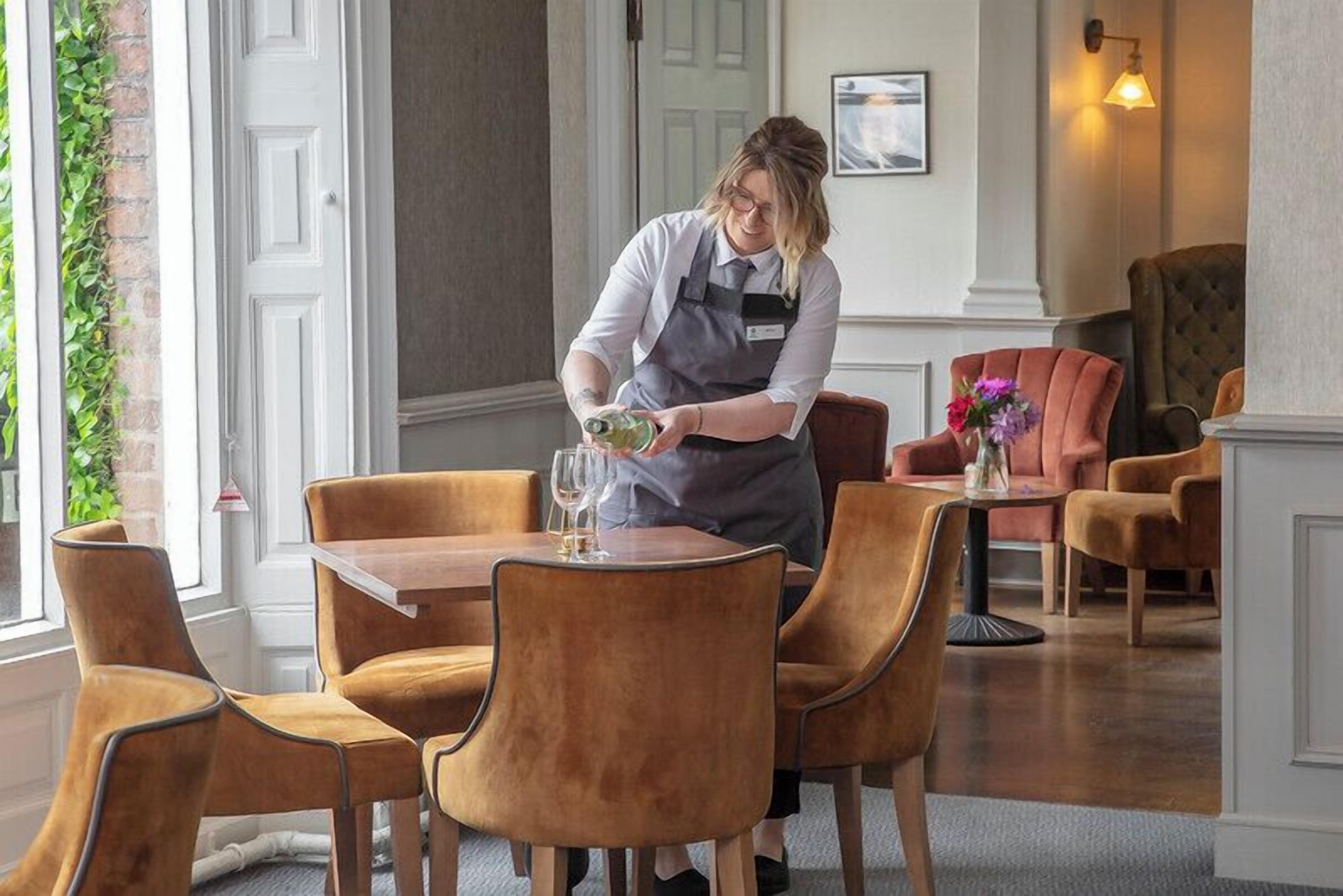 A person preparing a drink at a table in a room with a brick wall, a window, and a chandelier.