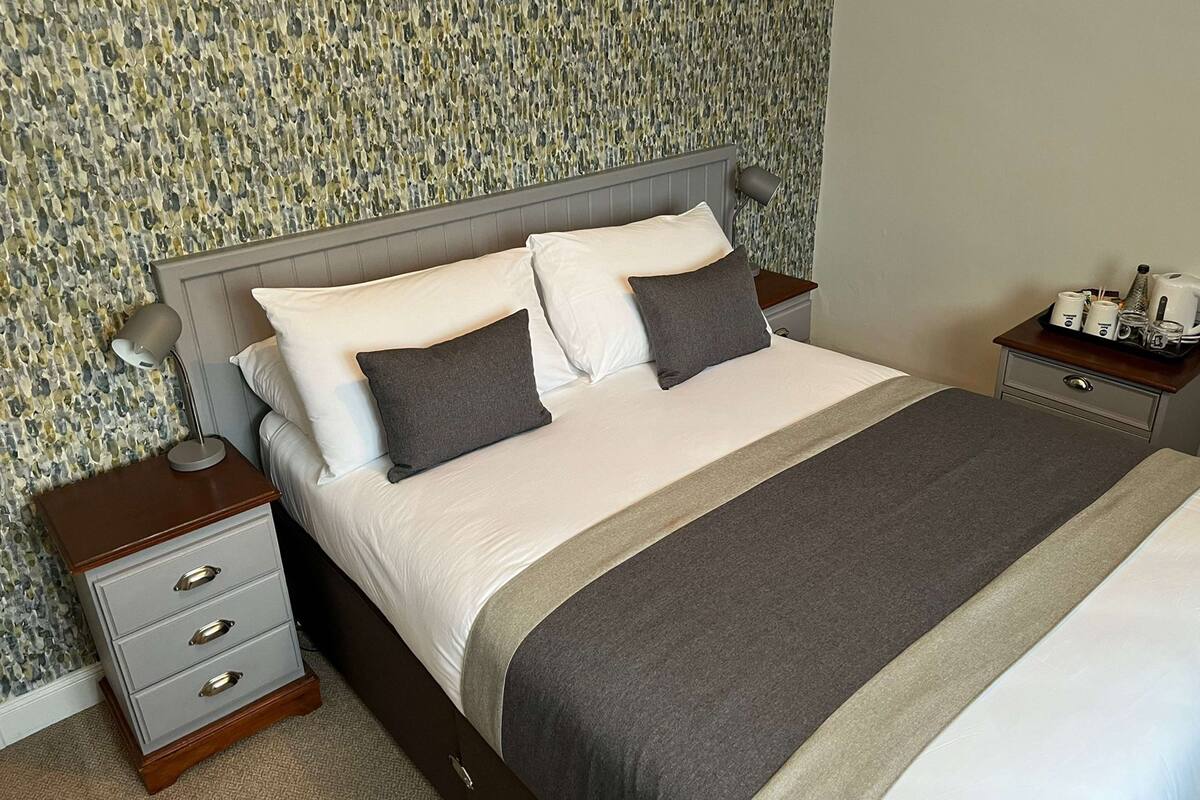 A neatly made bed with a grey and white color scheme, a bedside table with a lamp, and a tray with cups and a kettle.