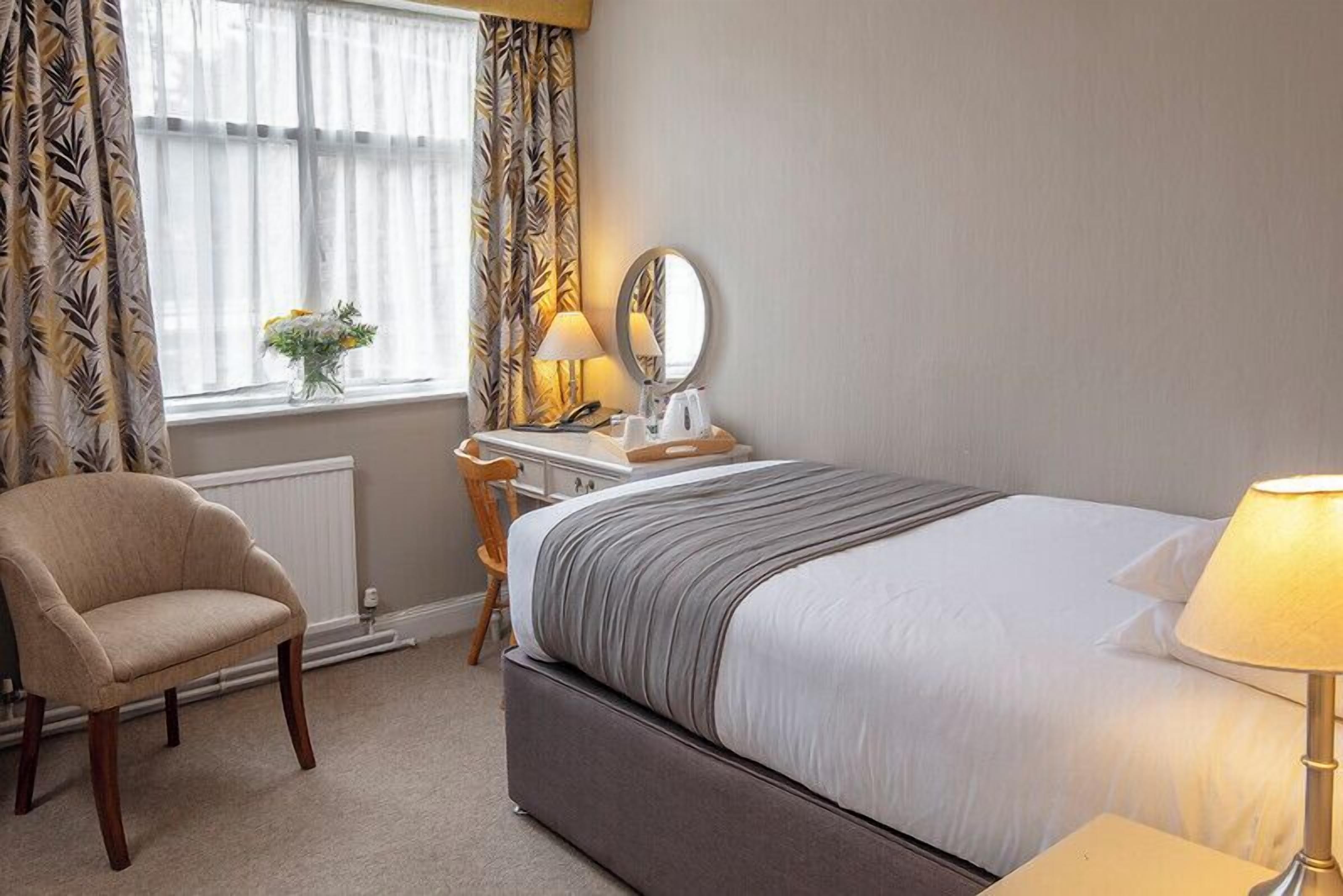 A hotel room with a bed, a chair, a desk with a lamp, a mirror, and a window with curtains.