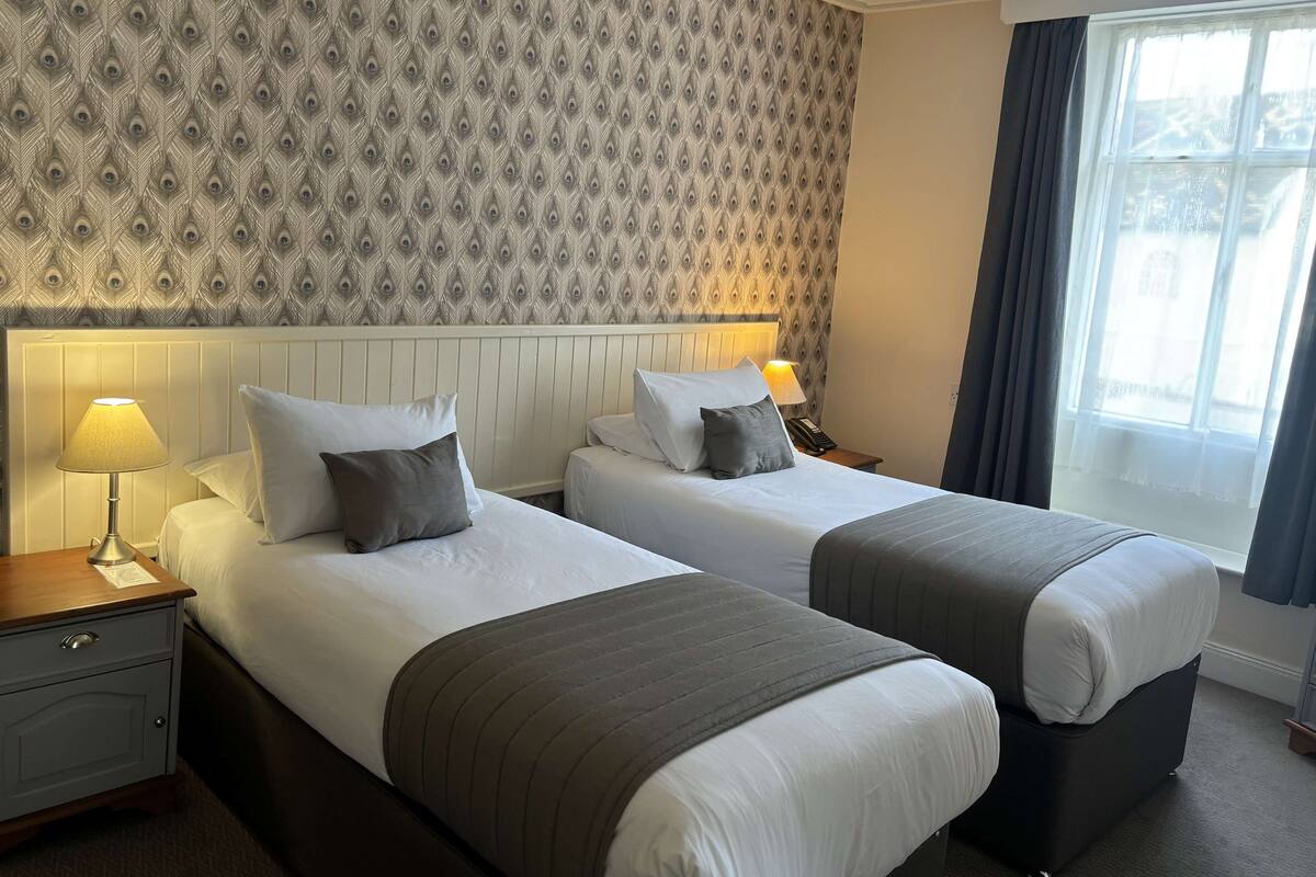 A hotel room with two beds, a nightstand, a lamp, a telephone, and a window with curtains.