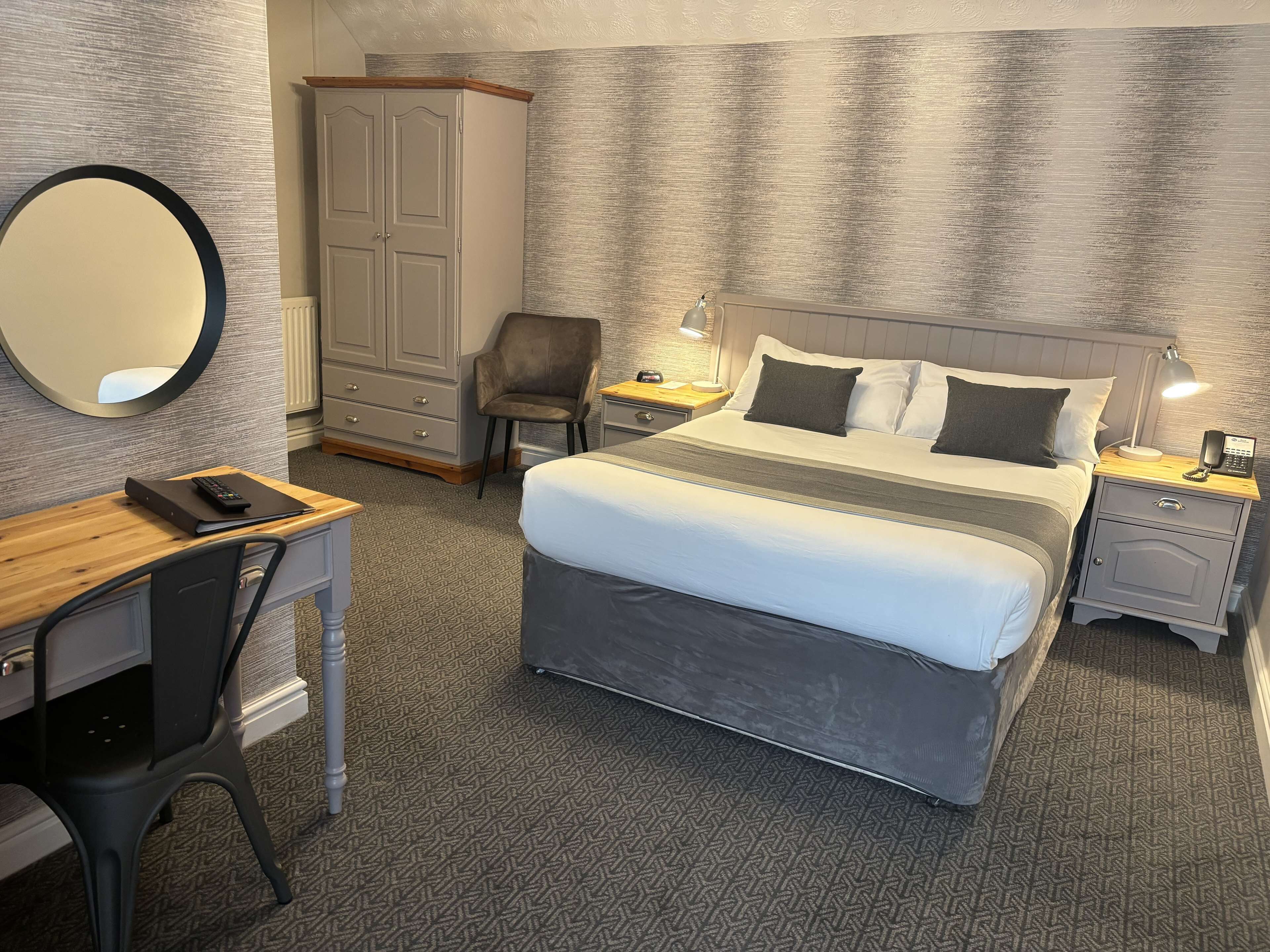 Et hotelværelse med seng, skrivebord, stol, garderobe, spejl og et natbord med lampe og telefon.