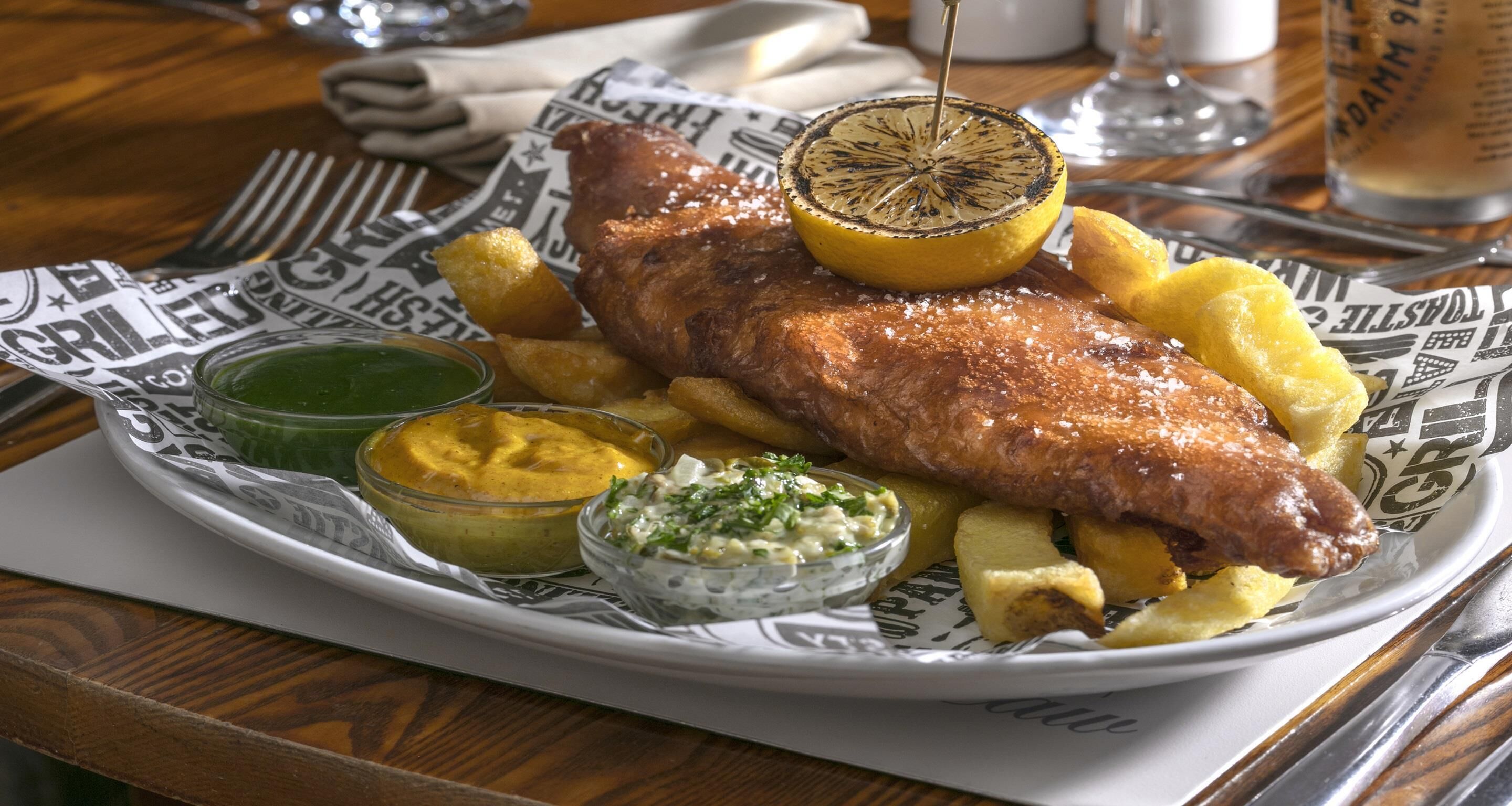 Un plato de pescado frito con patatas y rodajas de limón, acompañado de dos salsas para mojar.