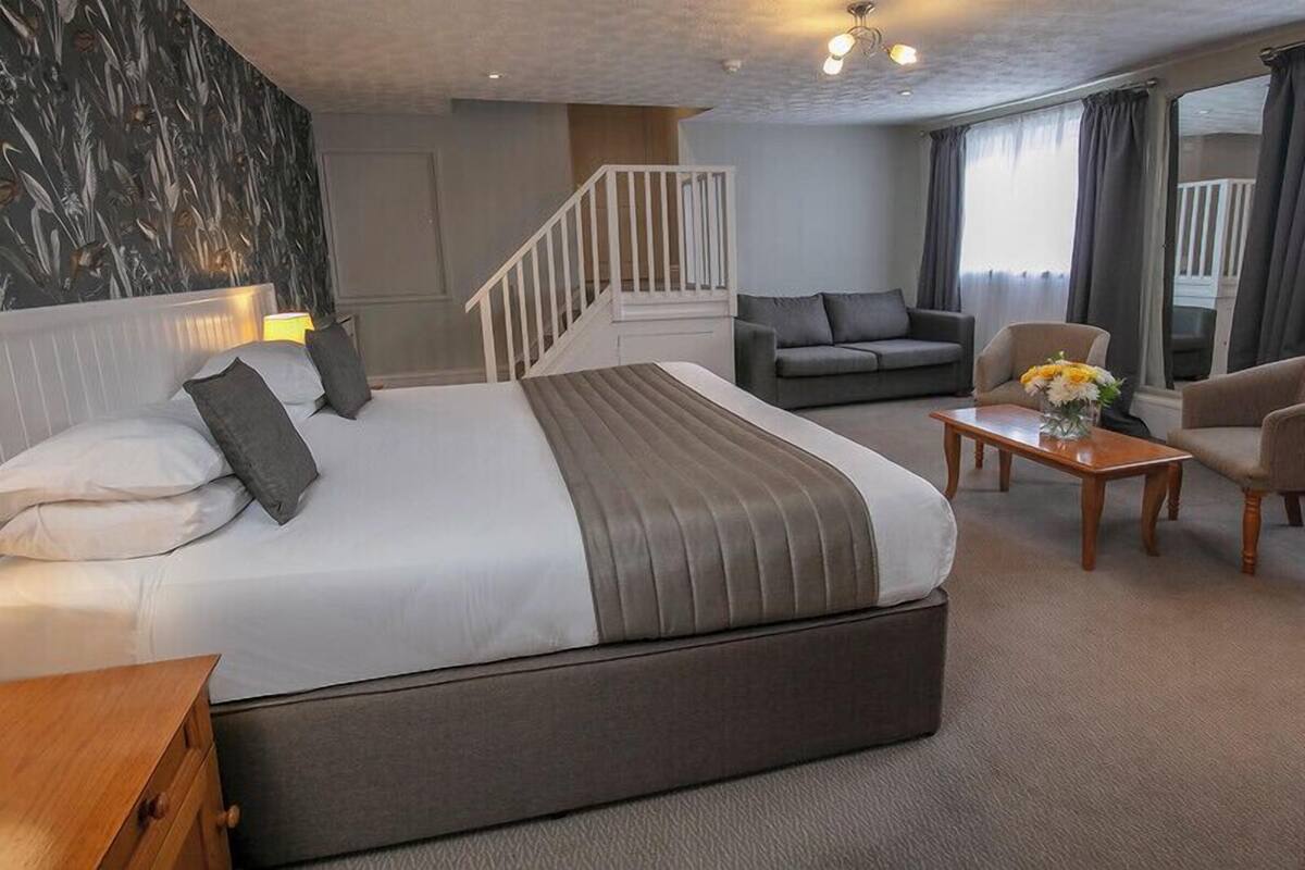 A modern hotel room with a large bed, a sofa, a coffee table, and a staircase.
