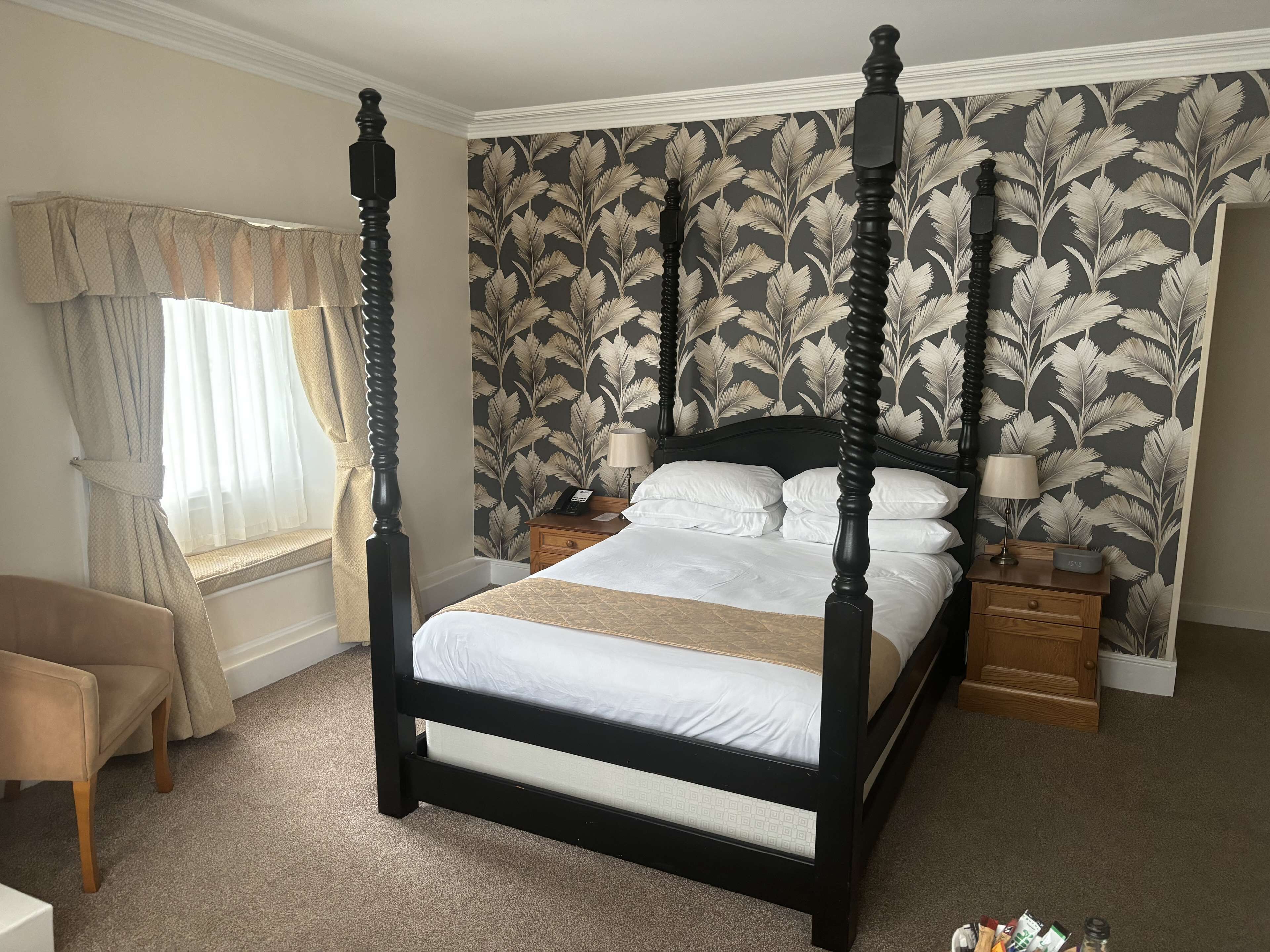 A four-poster bed with a dark frame, white bedding, and a floral wallpaper pattern. There are two bedside tables with lamps and a small bedside table with a clock.